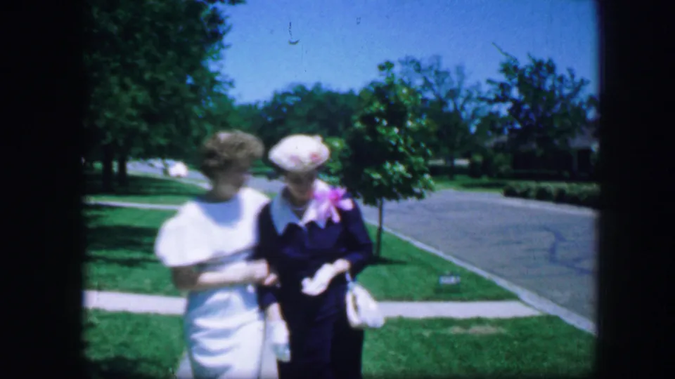 Two women in dressy 1960s clothing walk close together in a Fort Worth park, with the handheld 8mm frame showing lawn, trees, sidewalk, and road.