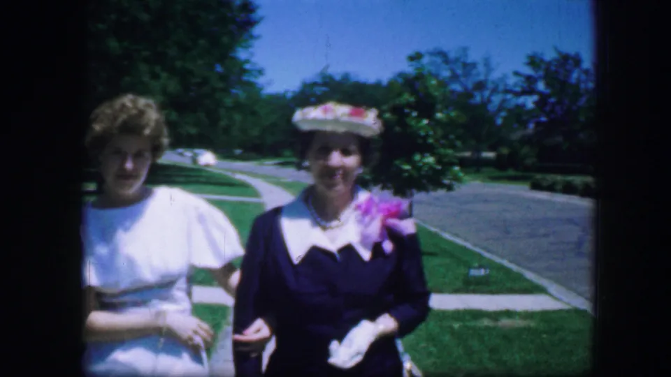 Close Fort Worth park frame of two adult women from 1964 home-movie footage, one in white and one smiling in a dark dress, gloves, and floral hat.