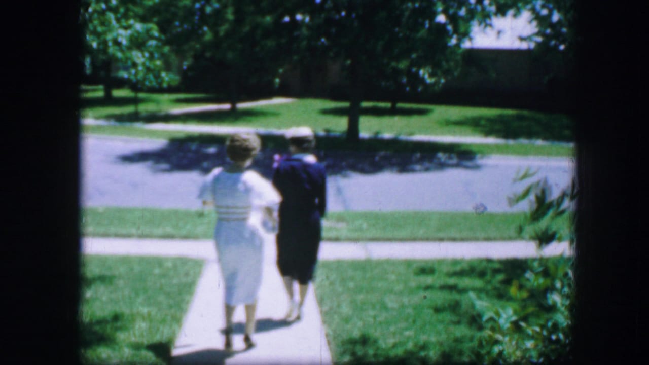 Two women walk away along a sidewalk toward a residential street.