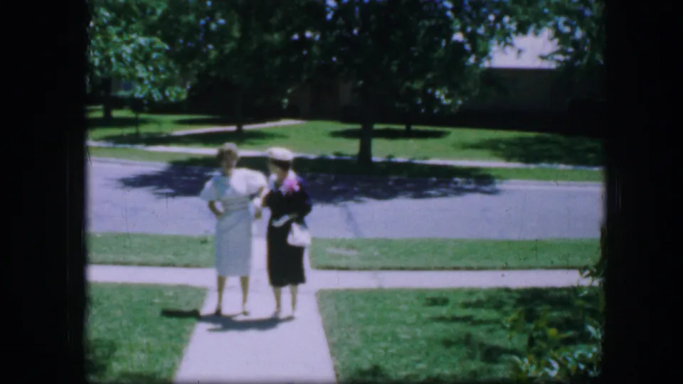 Two dressed women stand together on a Fort Worth sidewalk in 1964, framed by lawn, trees, and a residential street behind them.
