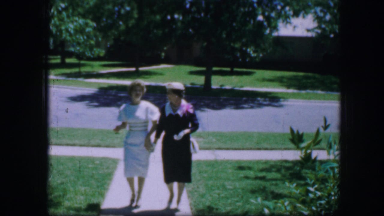 Two women walk closer to the camera, holding hands and carrying purses.