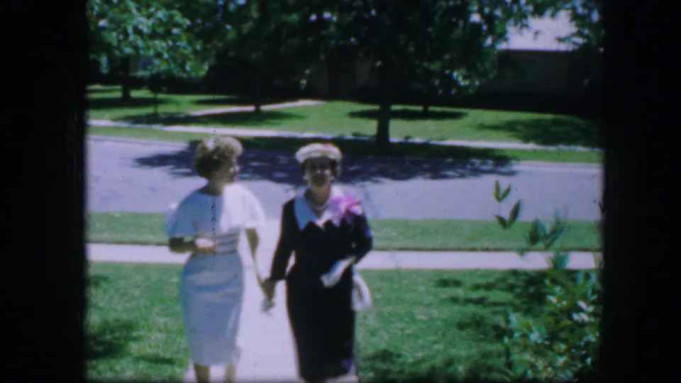 Two women in 1960s formal attire walk side by side on a sunny Fort Worth residential sidewalk.