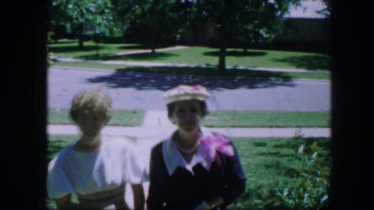 Two people in 1964 Fort Worth home-movie footage stand near a lawn and residential street, one wearing a hat and carrying a pink bouquet.