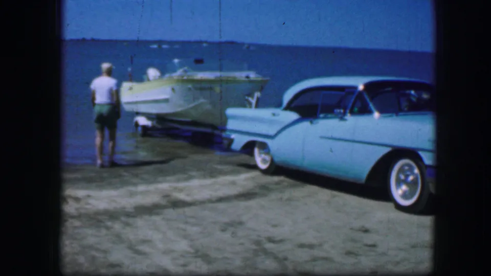 A man stands near a trailered boat and turquoise car beside a lake in 1964 Fort Worth home-movie footage.