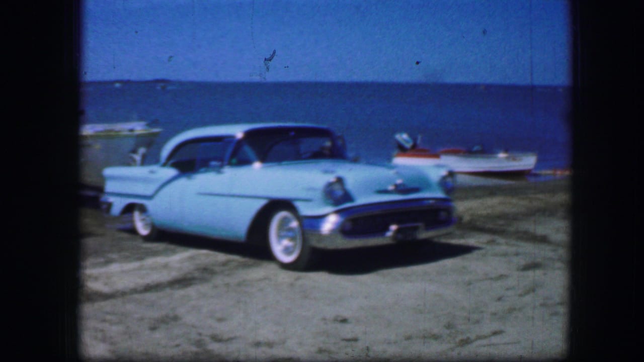 Light blue mid-century sedan with whitewall tires parked on sandy ground near blue water and small boats.