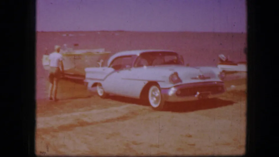 Faded 8mm frame of a pale classic sedan by a shoreline area with small boats and a distant standing figure.