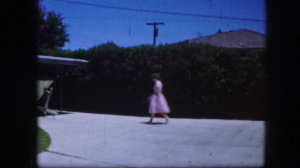 A woman in a pink dress walks across a paved area beside a tall hedge, with part of a vehicle visible at left.