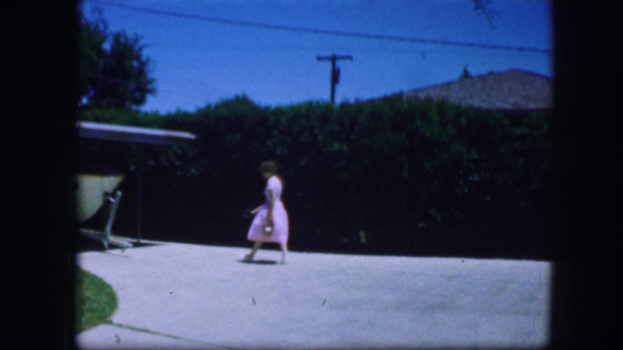 In 1964 Fort Worth home-movie footage, a woman in a light dress crosses a bright driveway near a hedge and covered structure.