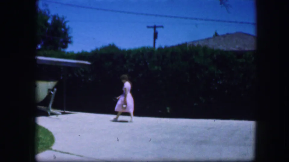 In 1964 Fort Worth home-movie footage, a woman in a light dress crosses a bright driveway near a hedge and covered structure.