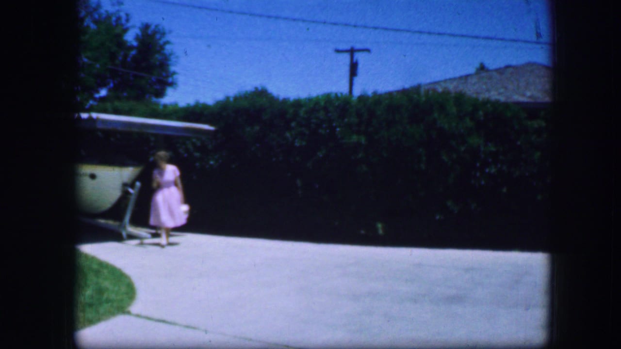 In Fort Worth, Texas, 1964 home-movie footage, a woman in a pink dress stands near a garage and driveway under a blue sky.