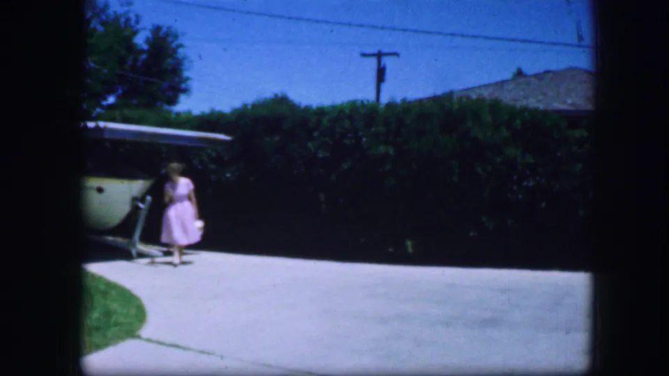 In Fort Worth, Texas, 1964 home-movie footage, a woman in a pink dress stands near a garage and driveway under a blue sky.