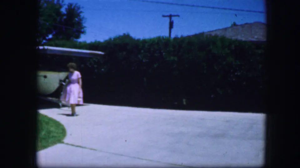 The woman in a pink dress walks from the garage area across a Fort Worth driveway in this 1964 8mm home-movie frame.