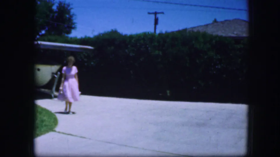 A woman in a pink dress walks toward the camera on a sunlit residential driveway in Fort Worth, Texas, in 1964.