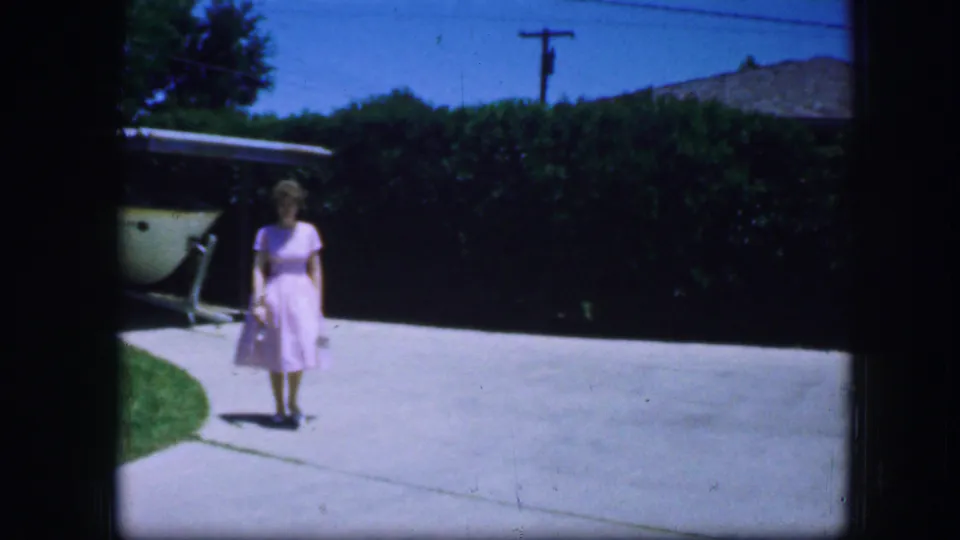 The woman in a pink dress stands or walks near the center of the driveway, framed by a hedge and garage in 1964 Fort Worth footage.