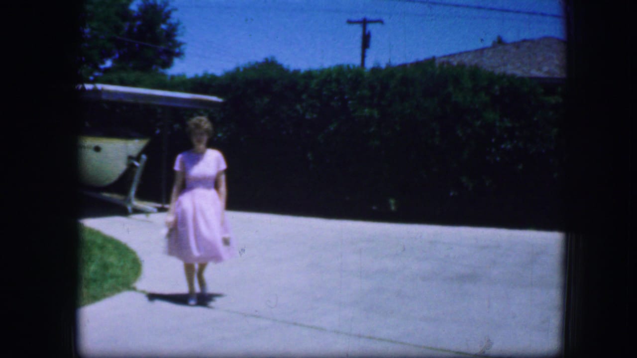 In 1964 Fort Worth home-movie footage, a woman in a pink dress walks toward the camera across a sunny suburban driveway.