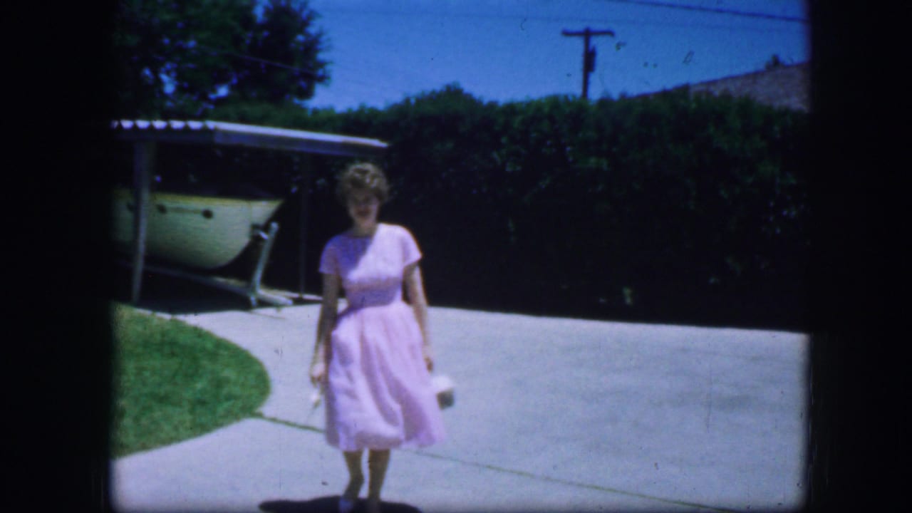 Woman in a pink dress closer to the camera on a driveway with hedge and carport behind her.