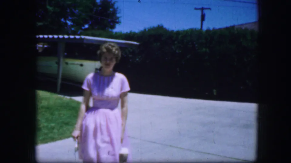 The woman nears the foreground in the 1964 Fort Worth driveway footage, holding a small light-colored item as she walks.