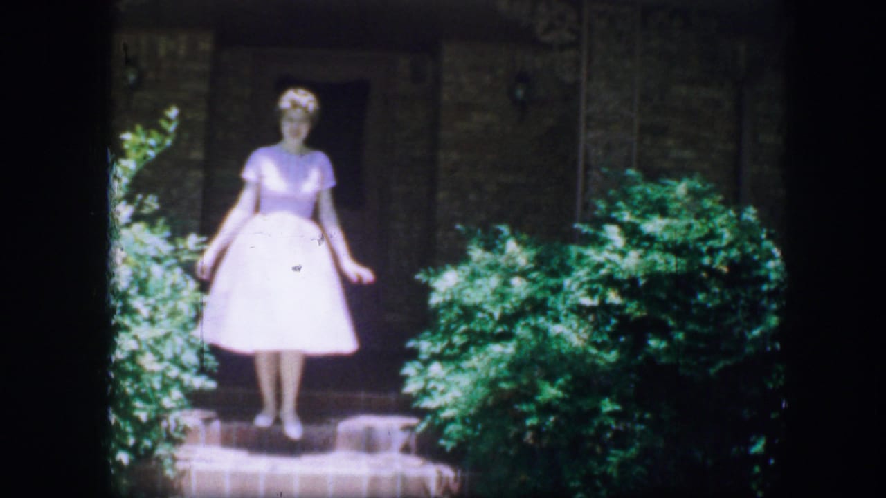 Young woman in a white dress stands on a stone garden path near greenery in Fort Worth, Texas, 1964 home-movie footage.
