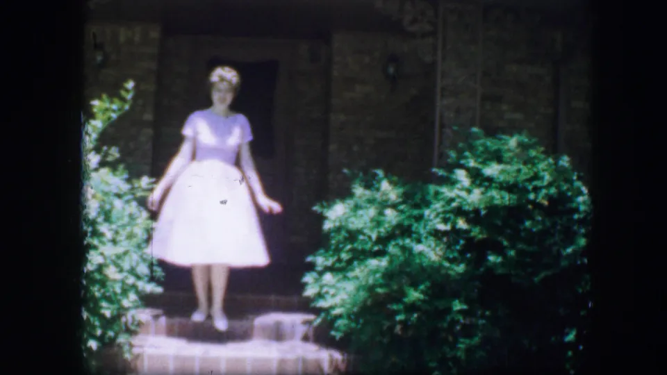 Young woman in a white dress stands on a stone garden path near greenery in Fort Worth, Texas, 1964 home-movie footage.