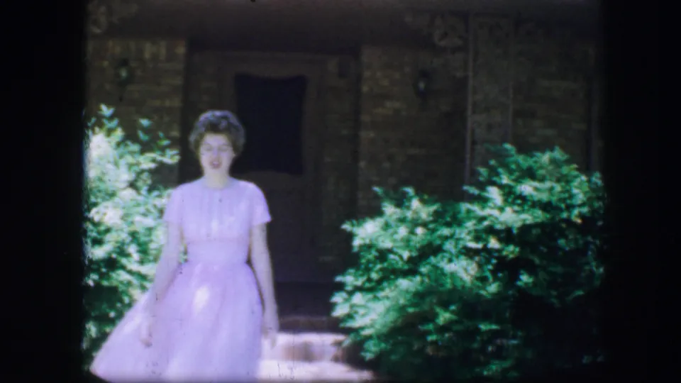 The woman approaches the camera in a pale dress, with sunlit shrubs and a dark building facade behind her in Fort Worth, 1964.