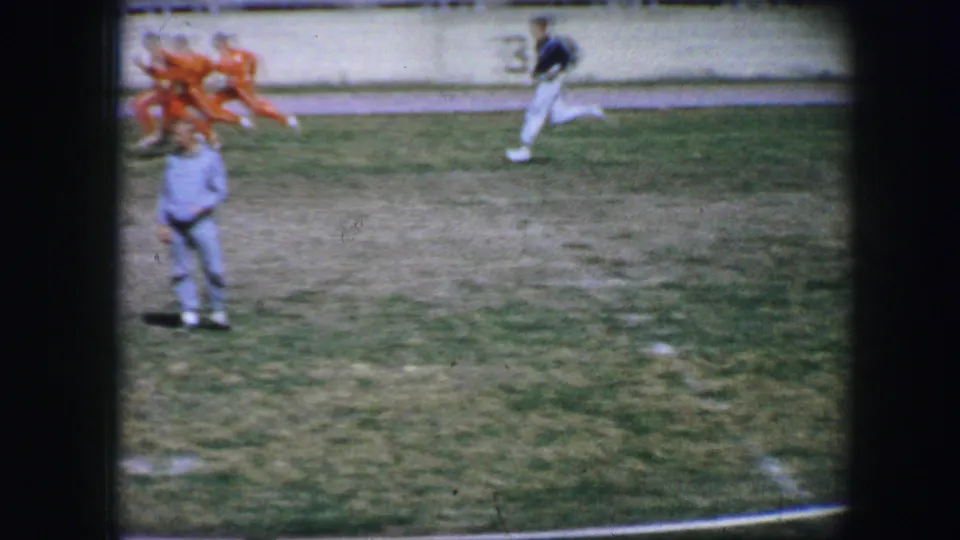 Fort Worth 1964 home-movie frame showing athletes moving across a grassy track field while an official stands nearby.