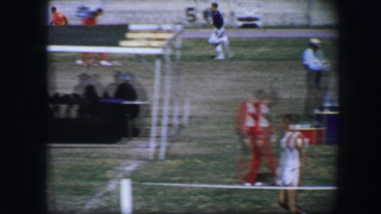 Athletes and officials near field markings with benches and spectators in the background.