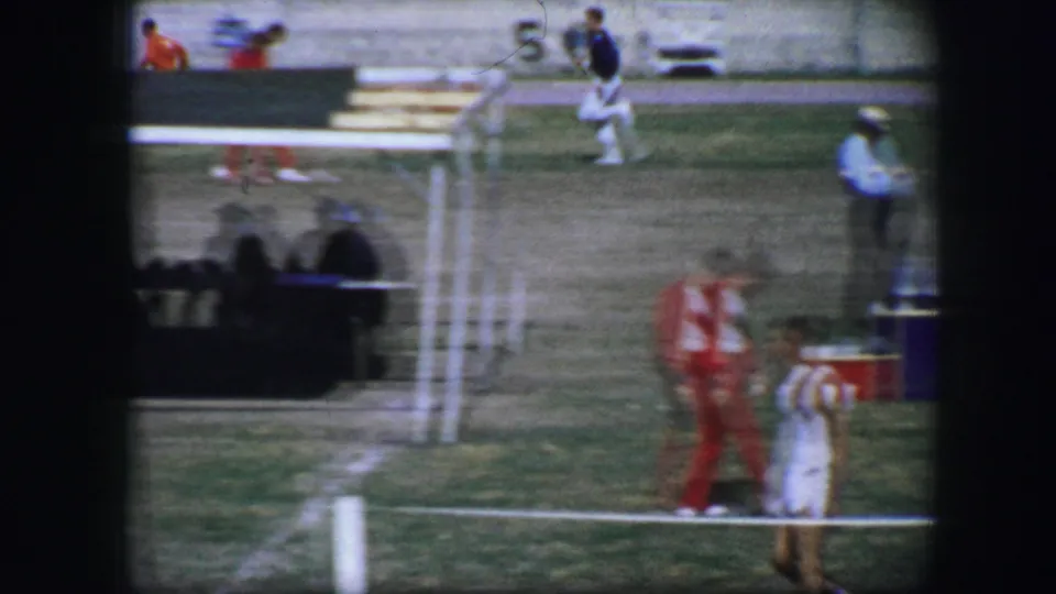 Fort Worth 1964 frame with athletes and officials spread across a grassy track area near benches and seated spectators.