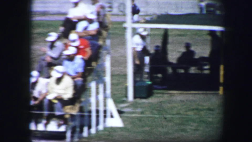 Fort Worth 1964 home-movie frame showing spectators seated near white railings beside the outdoor athletic field.