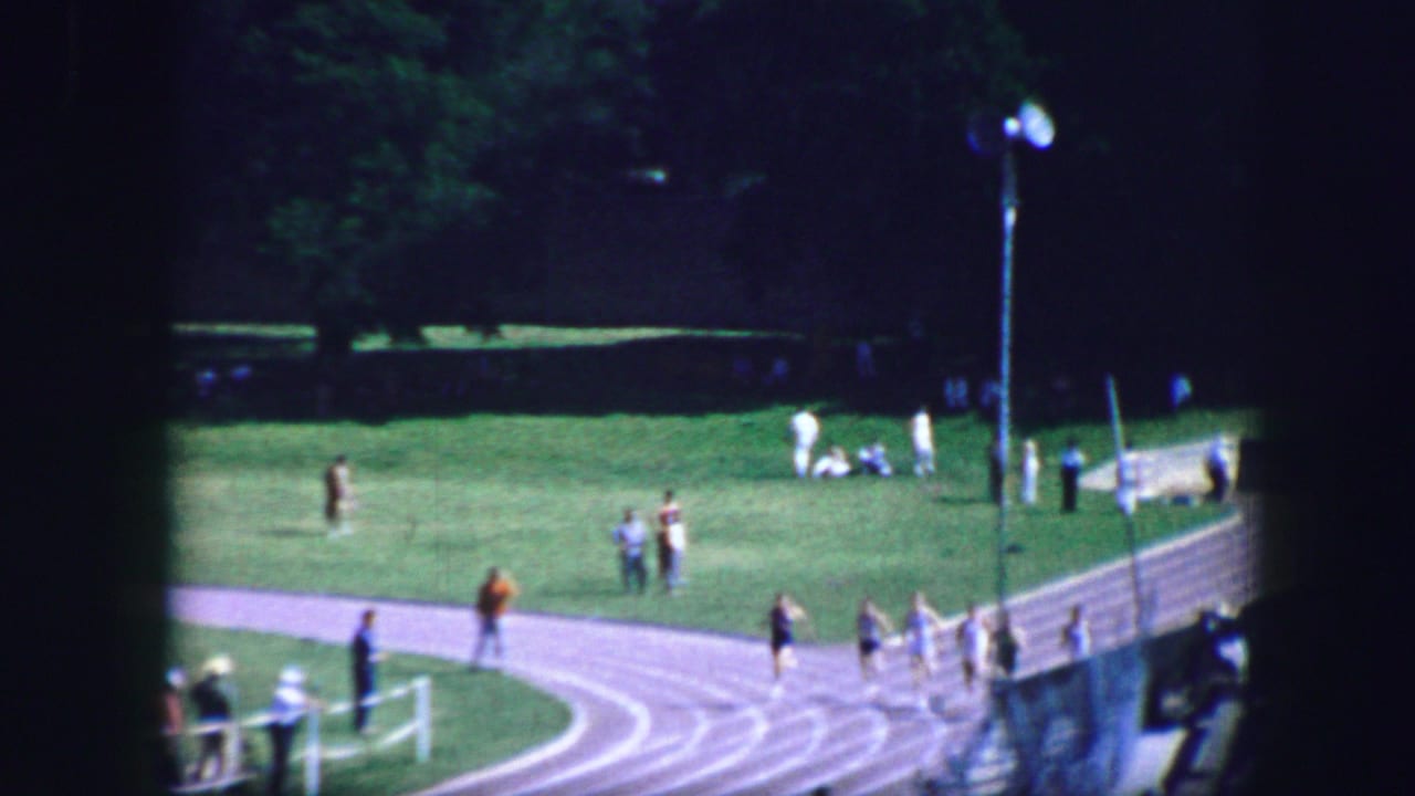 1964 Fort Worth home-movie view of runners on a curved outdoor track with spectators along the sideline.