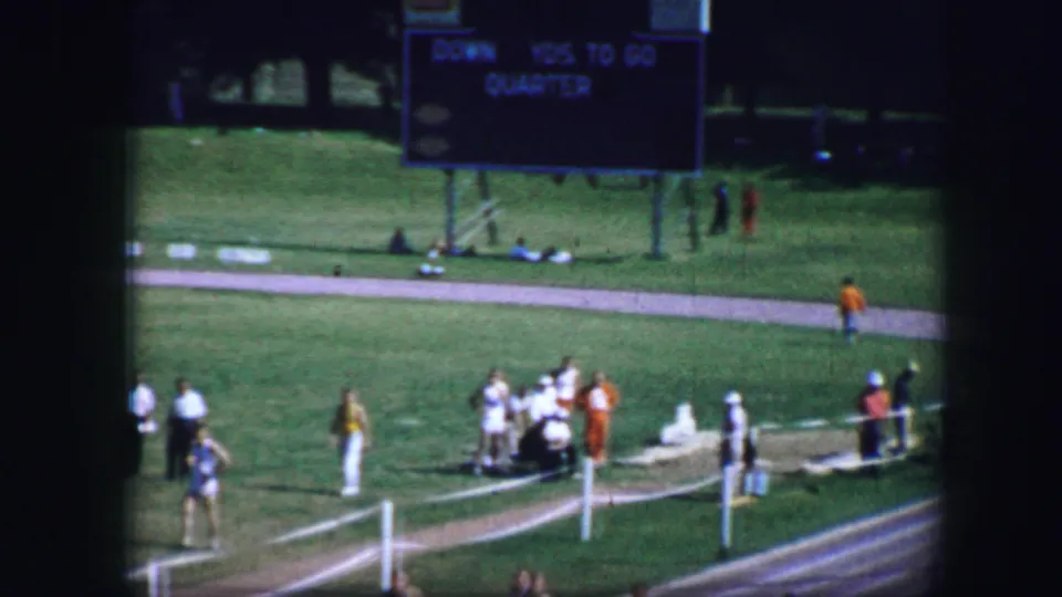 1964 Fort Worth home-movie frame with a scoreboard, sideline spectators, grass infield, and runners near the track edge.