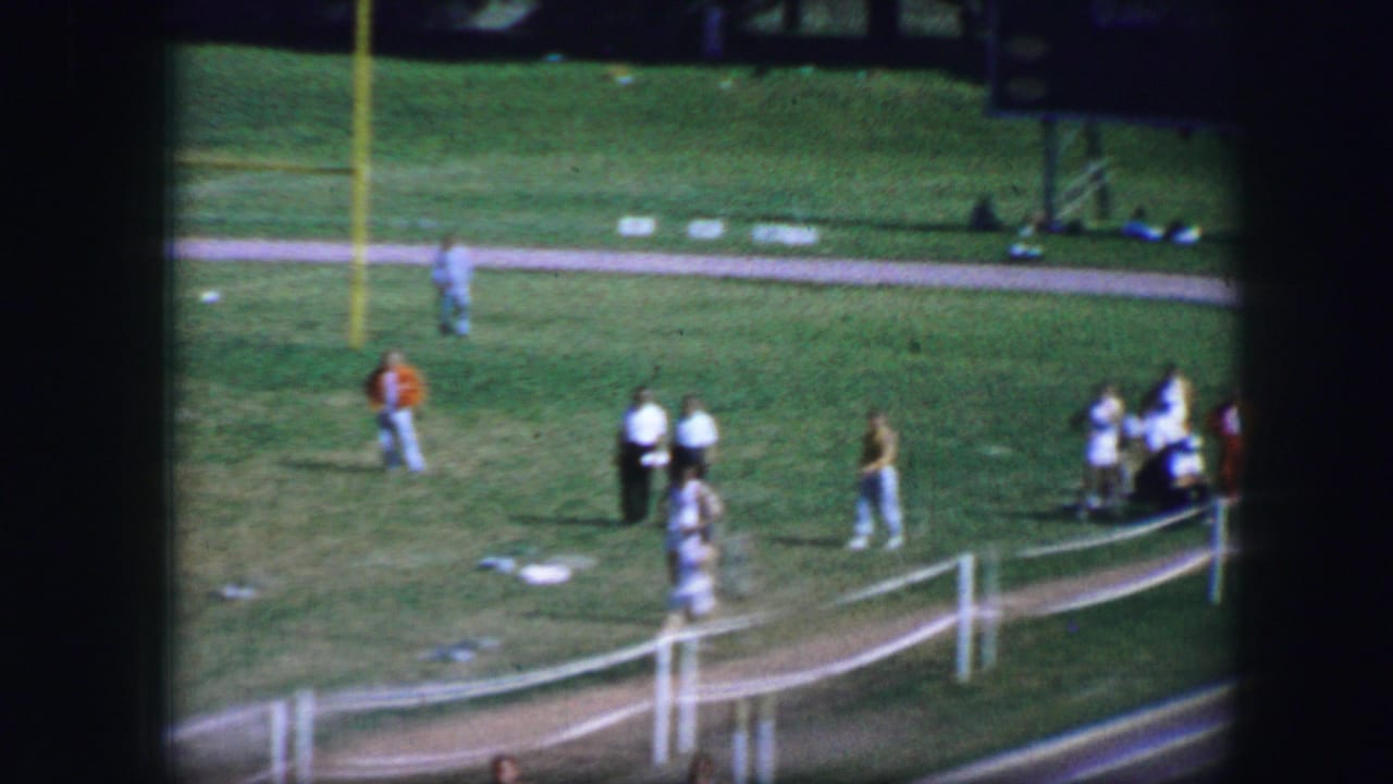 1964 Fort Worth home-movie frame showing spectators and athletes near a white fence beside a grassy race area.