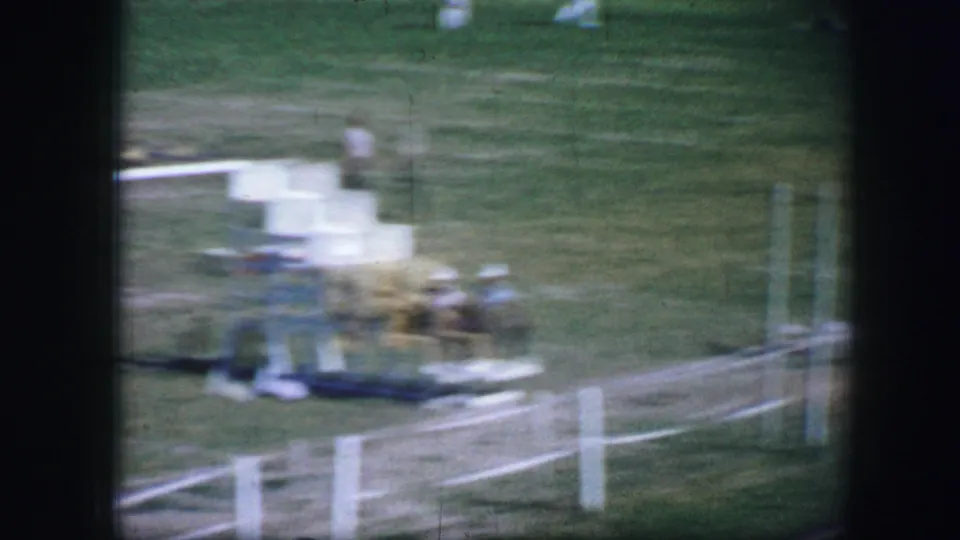 1964 Fort Worth frame with blurred figures gathered near a white fence and field equipment by the race area.