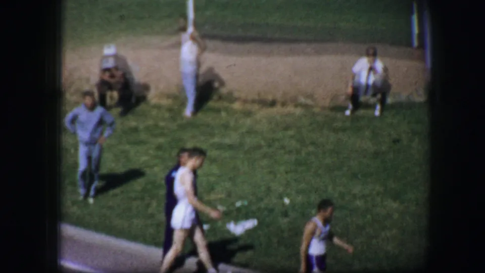 1964 Fort Worth frame showing track athletes walking near seated spectators beside the grassy competition area.