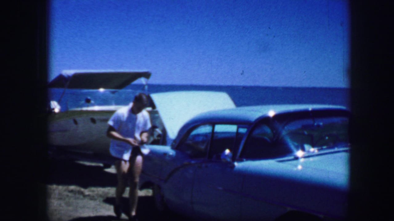 Young man in a white shirt stands near a light blue car with a boat behind him.