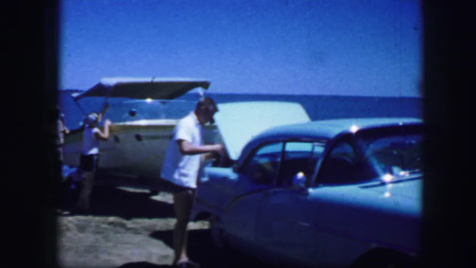 The man stands by the light blue car as another person appears at the left near the boat in this 1960s travel scene.