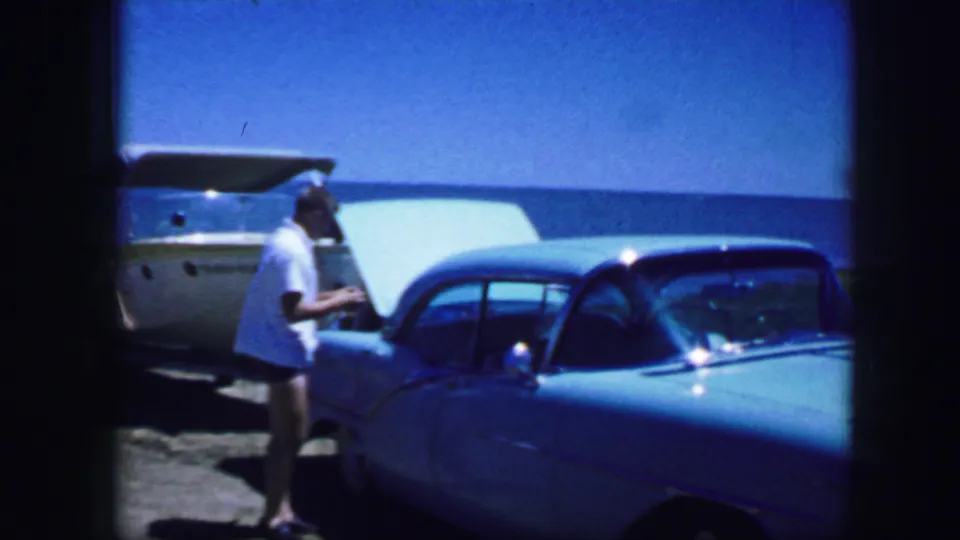 A man in a white shirt stands at the front of a light blue classic car, framed by a boat, sky, and beachlike ground.