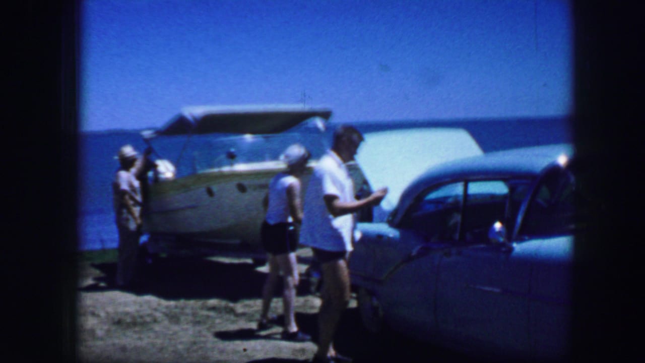 Several adults walk between a blue car and a light-colored trailer near open water.