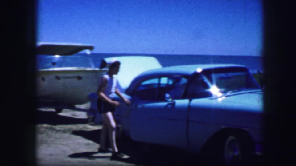 Fort Worth, Texas, 1964 home-movie frame showing a person beside a blue classic car near a pale trailer or boat under a clear sky.