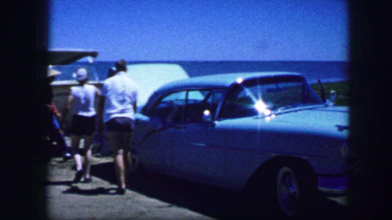 1964 8mm home-movie frame showing adults by a light blue mid-century car with an open trunk near a waterfront setting.