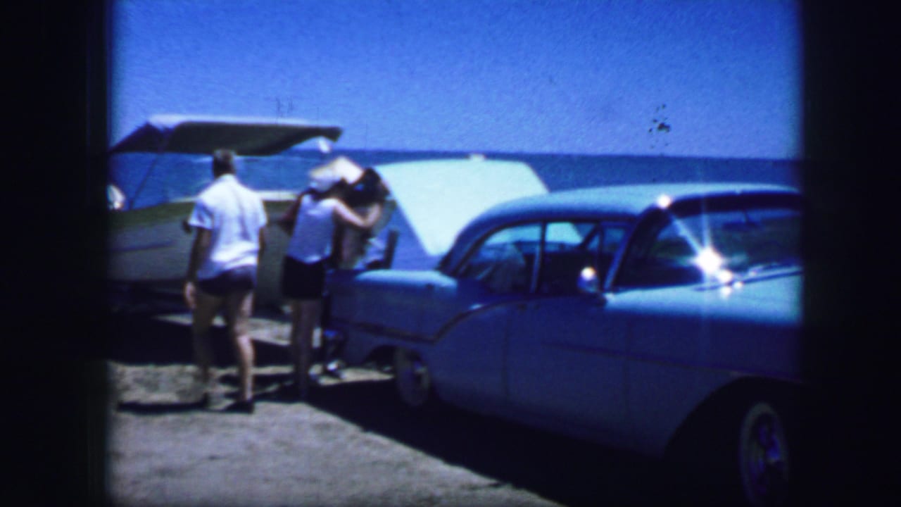 Two adults stand at the open rear of a car beside a light blue sedan near the water.