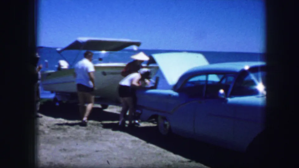 A 1964 8mm frame shows adults handling items near an open trunk, with a light blue mid-century car and waterfront behind them.