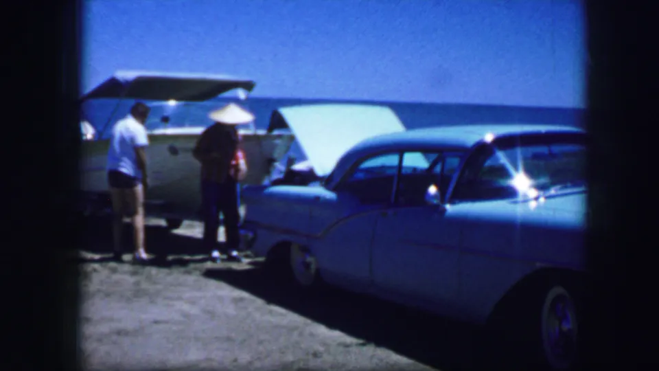 Two adults pause beside parked mid-century cars, including an open trunk, in waterfront 1964 8mm home-movie footage.