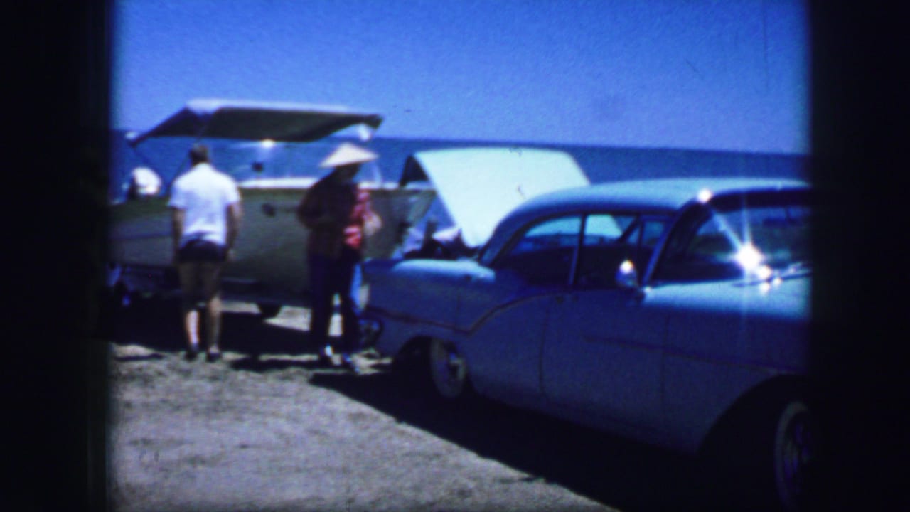Men stand beside a blue sedan and small boat near open water under a bright sky.
