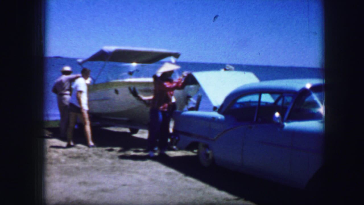 Several men stand near a small boat and blue car parked beside the water.
