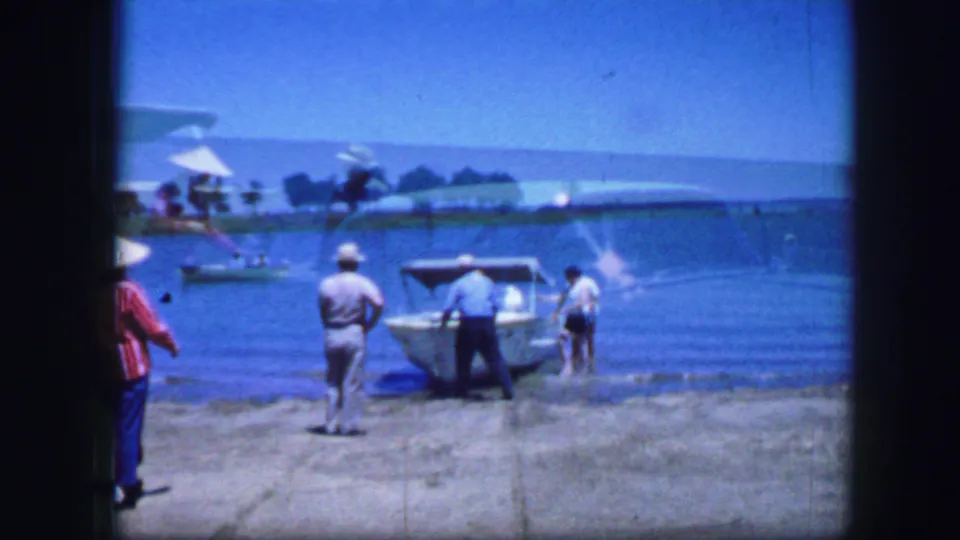 Home-movie frame shows men moving a small boat into shallow water, with a bystander partly visible at the left edge.