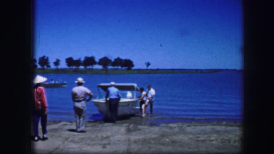 1964 Fort Worth-area home-movie frame showing men around a small boat at the shoreline, with open water and a distant tree line.