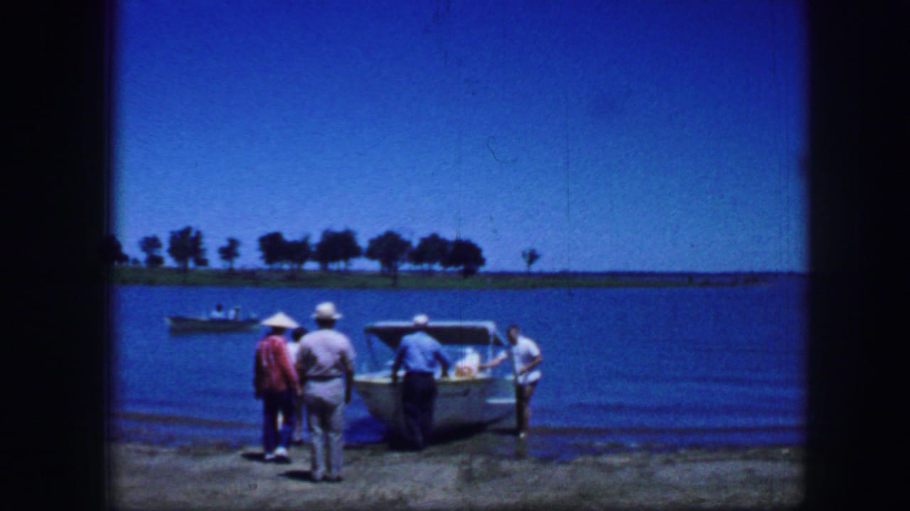 Adult men in hats stand beside a small boat at a concrete dock with water and trees behind them.