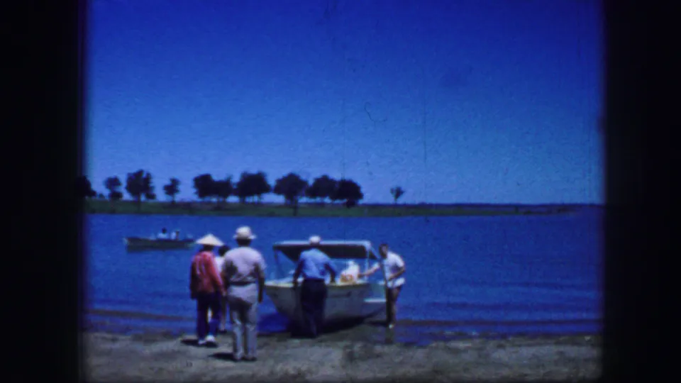 1964 Fort Worth home-movie frame showing adult men near a small boat on a concrete dock beside calm water.