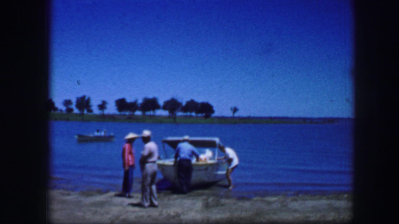 Four adult men stand around a small boat at the shoreline while another boat sits on the water.