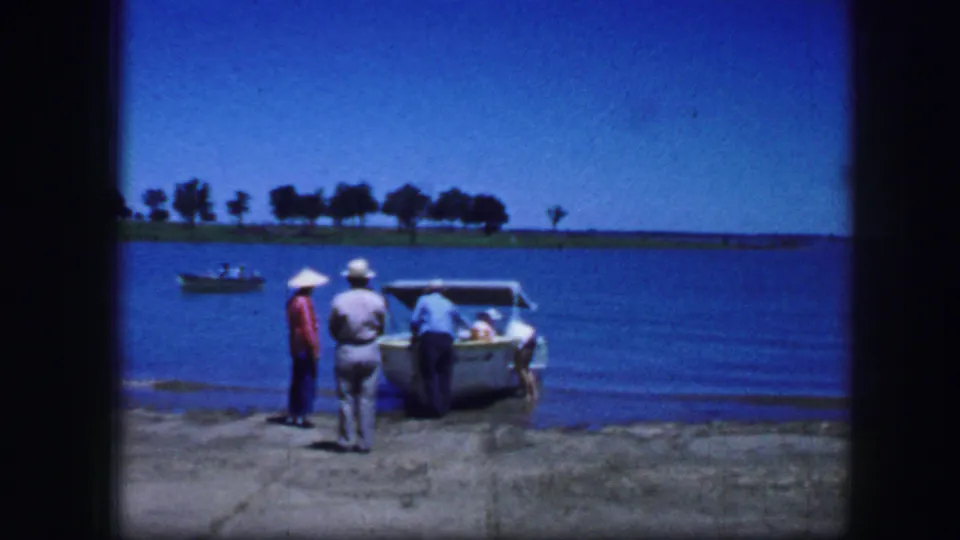1964 Fort Worth 8mm frame of adult men at a concrete dock beside a small boat, with calm water and trees beyond.